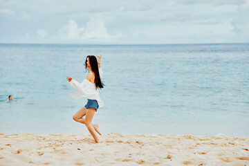 A woman runs along the beach in summer clothes on the sand in a yellow T-shirt and denim shorts white shirt flying hair ocean view