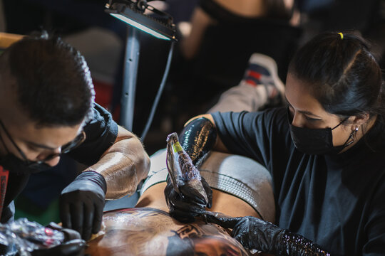 top view of male and female tattoo artists working simultaneously on the back of a client, use of biosafety elements