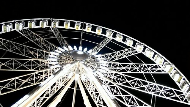 large white metal ferris wheel with small cabin heap, giant attraction illuminated with bright led light on dark black sky, modern entertainment diversity