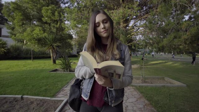 Mujer joven camina por el parque leyendo un libro durante el atardecer.