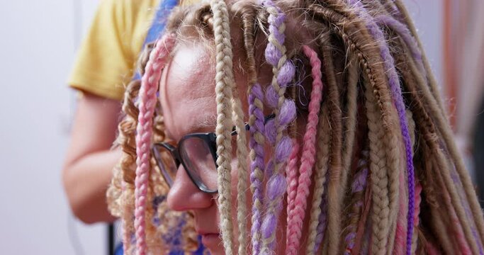 Tired master of beauty salon monotonously unweaves Afro braids client glasses end of shift. Close-up of creative bright hairstyle correction colored strands personality. Image change, unravel pigtails