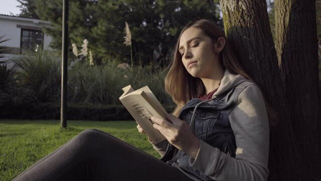 Mujer joven leyendo sentada y recargada en un arbol durante el atardecer.