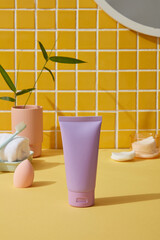 Makeup sponge, towel, cotton pads and plant pot on yellow bathroom background. Purple tube with no label for product advertising
