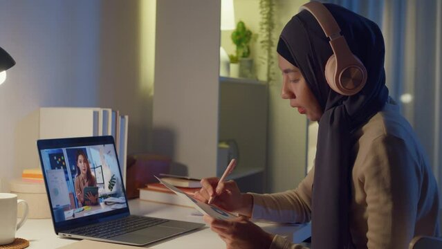 Young happy Asia muslim woman secretary wear hijab casual clothing video conference meeting on laptop computer with businesswoman at home overtime working at night. Online learning concept.