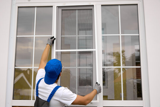 View from back master in working uniform of blue color, in his hands high raises mosquito net for installation on window. Professional installation of protective nets on windows of different sizes