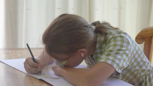 A little girl with glasses sitting at a table and drawing, low head tilt, incorrect posture, bent low, stooped, poor eyesight