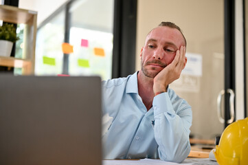 Bored and tired Caucasian male engineer sits at his desk, hand on chin, looking at his laptop screen
