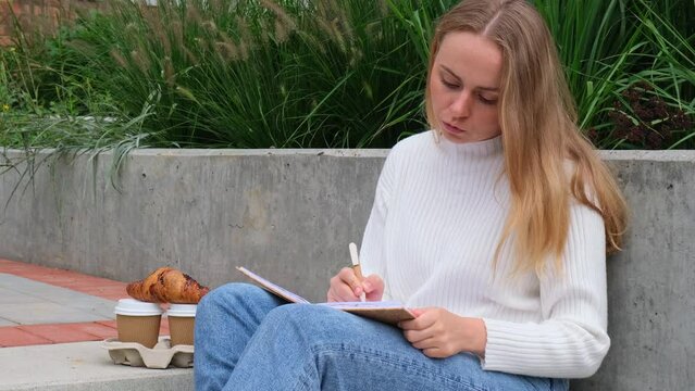 Young student study with notebook in park. Drinking coffee and eat croissants. Writing gratitude journal self reflection self discovery Outdoors warm autumn seashore. 