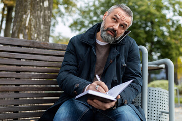 Fototapeta premium mature adult man talking on the phone making notes in a diary while sitting in the park