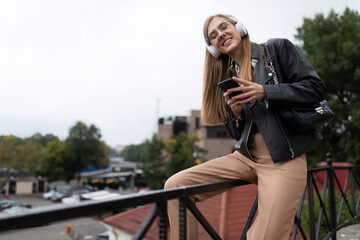 young adult woman with a wide smile on her face listens to music in headphones while sitting on the parapet with a phone in her hands