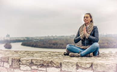 Fototapeta na wymiar Beautiful young brunette woman with long hair meditating, wearing warm winter clothes, sitting on a stone parapet by the lake. Toned