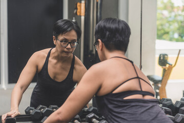 Fototapeta premium A fit middle aged asian woman with short hair getting into the zone before a workout. Picking up a dumbbell from the rack. Looking at her reflection in the gym.