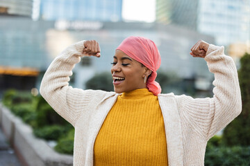 Latina woman, fighting breast cancer, wears a pink scarf, and clenches her arms as a survivor...