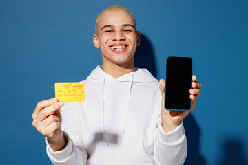 Young man of African American ethnicity wear white hoody use blank screen mobile cell phone credit bank card shopping online order delivery book tour isolated on plain dark royal navy blue background.
