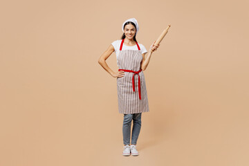 Full body young smiling happy housewife housekeeper chef baker latin woman wear striped apron toque hat hold rolling pink look camera isolated on plain pastel light beige background Cook food concept
