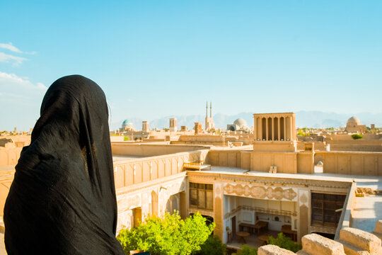 Yazd, iran - June 3rd, 2022: Iranian muslim woman wearing abaya in rooftop cafe enjoy sightseeing rooftops panorama with wind-catchers in YAzd. Famous travel Iran destination