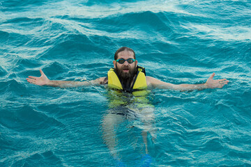 A bearded man diver in a life jacket swims in clear azure sea water.