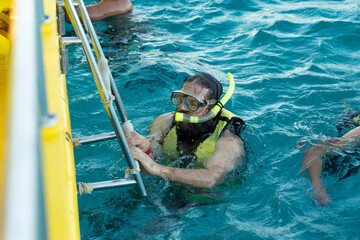 A bearded man diver in a life jacket climbs on board a boat in the sea.