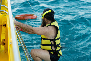 A bearded man diver in a life jacket climbs on board a boat in the sea.