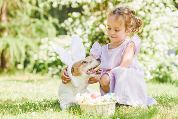 Cute little girl hugging her pet dog wearing Easter bunny ears costume. Basket with decorated eggs and flowers.