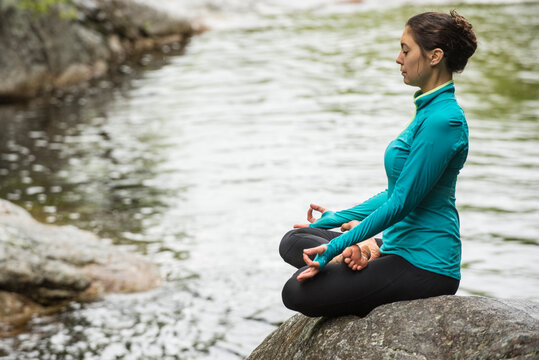 Woman meditating in lotus position while practicing yoga on rock in river, Gorham, New Hampshire, USA