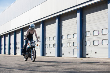 Woman riding on her vintage motorcycle wearing a reflective helmut face shield with a line of auto garage in the background.