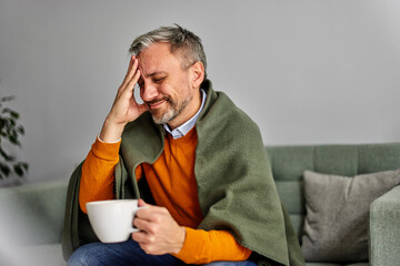 A sick senior man is feeling a headache migraine, drinking tea, and sitting on the couch.