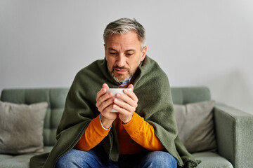 A sick senior man warming his hands with a cup of tea, wrapped in a blanket at home.