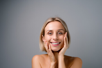 Skin care. Blonde woman with beauty face touching healthy facial skin. No makeup. Grey background.