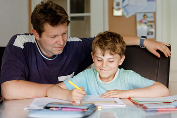 Cute little school kid boy at home making homework with dad. Little child writing with colorful pencils, father helping him, indoors. Elementary school and education.