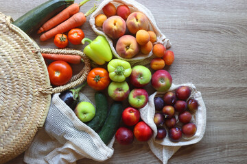 Straw bag and reusable fabric bags filled with various healthy fruit and vegetables. Wooden background, top view.