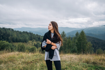 An attractive woman in casual clothes stands in the mountains against a background of beautiful scenery and looks away with a serious face.