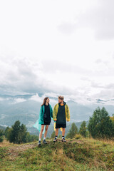 Fototapeta na wymiar A beautiful couple man and woman in casual clothes and raincoats in rainy weather are standing in the mountains on the top against the background of a cloudy beautiful landscape.