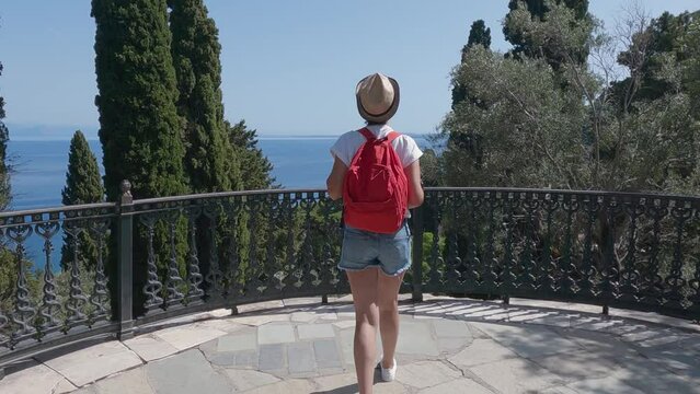 Tourist woman with hat and backpack enjoy the view from the Terrace in the Garden in Achilleion Palace, in Corfu island, Greece, slow motion