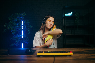 Fototapeta na wymiar Positive streamer girl in a headset plays online games on the computer and hugs a rubber chicken toy.