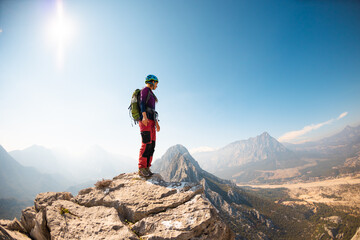 girl climber in a helmet and with a backpack stands on a mountain range against the backdrop of mountains and the sky. mountain climbing - obrazy, fototapety, plakaty
