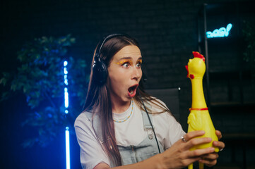 A shocked woman gamer in a headset holds a toy chicken in her hands and looks at it with a...