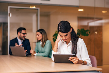 A tired businesswoman reading something on a tablet, two colleagues talking.