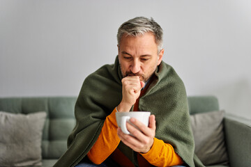A sick senior man coughs, feeling sick, holding a cup of tea, resting on the couch.