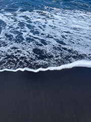 Gentle wave of white foam rolls over a black sandy beach.