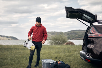 Windsurfer and camper unpacking equipment from a car in nature near the lake shore.