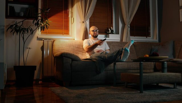 Bearded Caucasian young happy man wearing glasses is sitting on sofa in cozy evening room and watching tv.