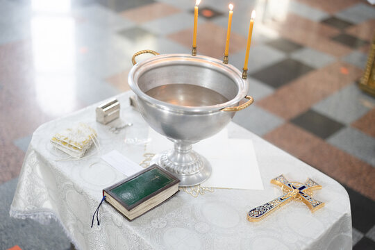 Baptismal font with wax candles and cross are on the table near Holy Bible.