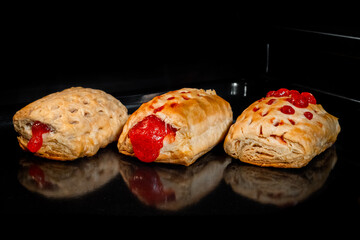Three baked puff pastry buns with red strawberry jam on tray in electric oven, black background. Homemade bakery, food, cooking and pastry concept