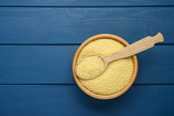 Bowl and spoon with raw couscous on blue wooden table, top view. Space for text