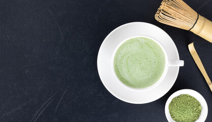 Traditional matcha green tea latte in white cup, bamboo whisk and matcha powder on black table. Top view. Copy space.