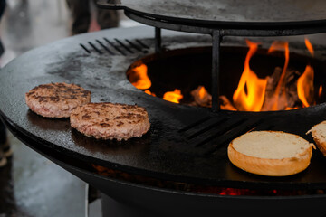 Process of grilling fresh meat cutlets and buns for burgers on brazier with hot flame at summer local food market - close up view. Outdoor cooking, gastronomy, cookery, street food concept