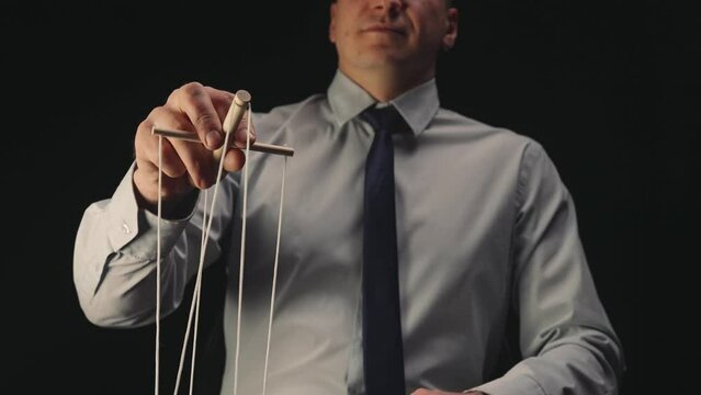 A businessman in a white shirt and black tie controls a puppet with a wooden manipulator and strings. A puppeteer controls a doll marionette on a black isolated background. Close up.