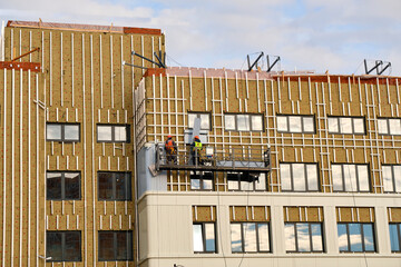 Builders insulate walls of facade of multi-storey residential building with mineral insulation, while on construction cradle. Heat saving concept.