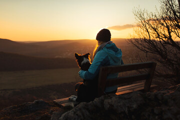 border collie puppy dog and young woman owner sitting on a bench on a mountain top at sunset seen...